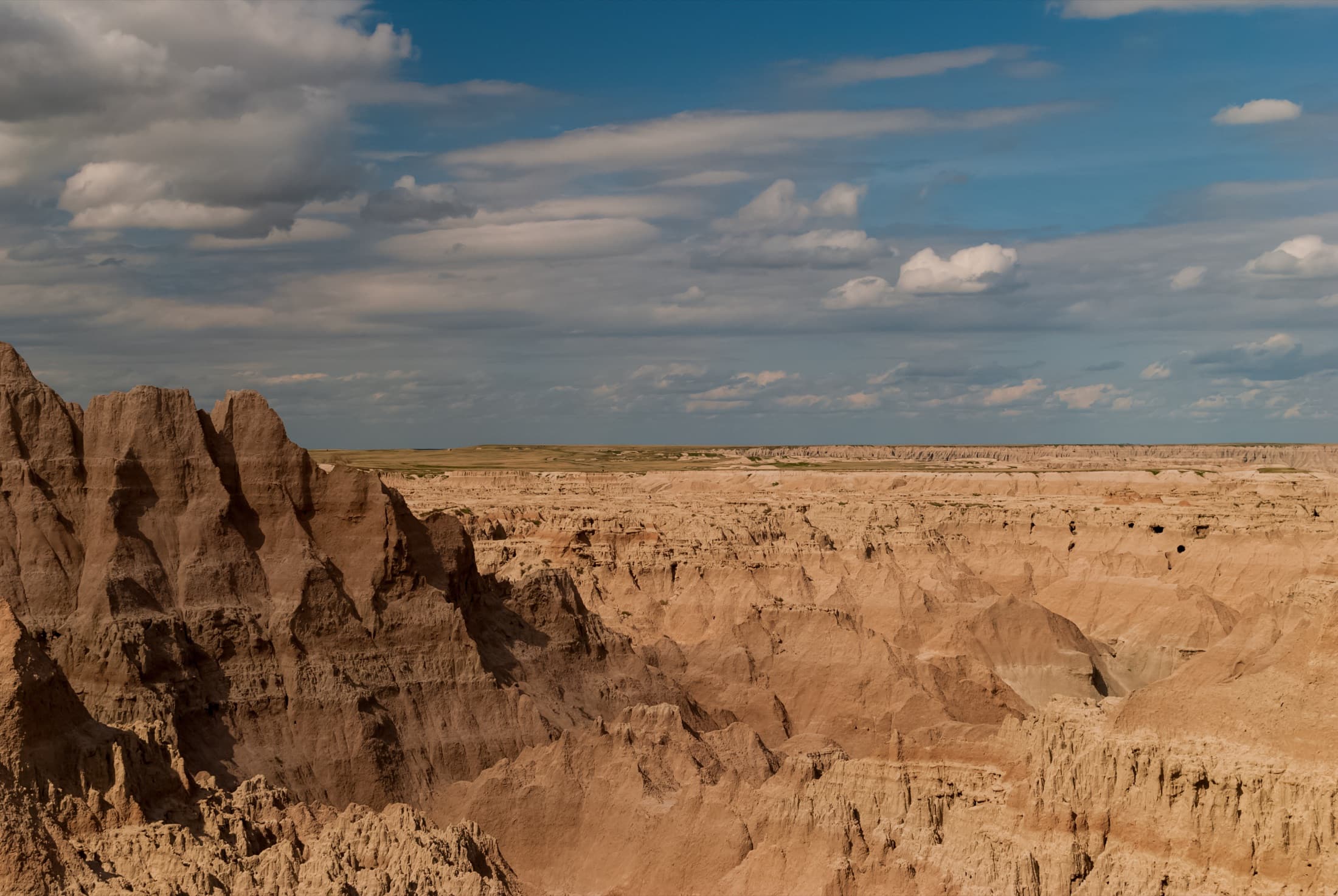 Wide panorama of the eroded buttes, layered spires, and sun-bleached clay formations of Badlands National Park, South Dakota, sweeping out toward mixed-grass prairie under a broad sky.