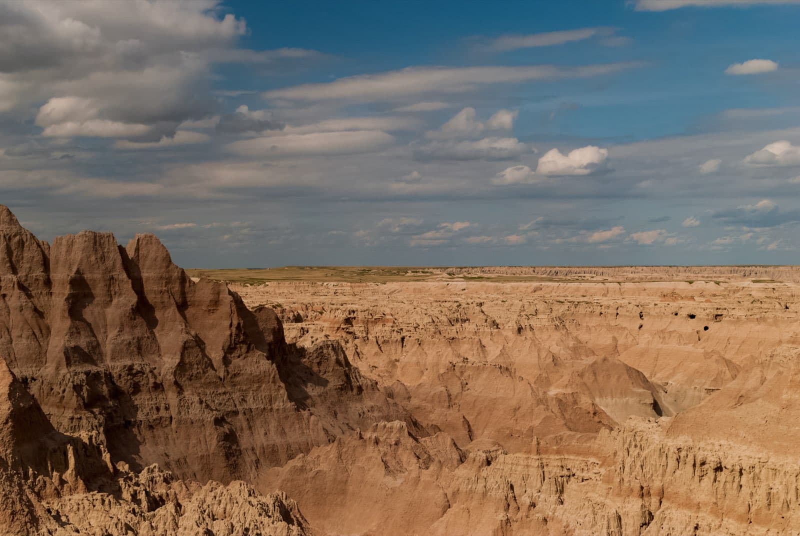 Wide panorama of the eroded buttes, layered spires, and sun-bleached clay formations of Badlands National Park, South Dakota, sweeping out toward mixed-grass prairie under a broad sky.