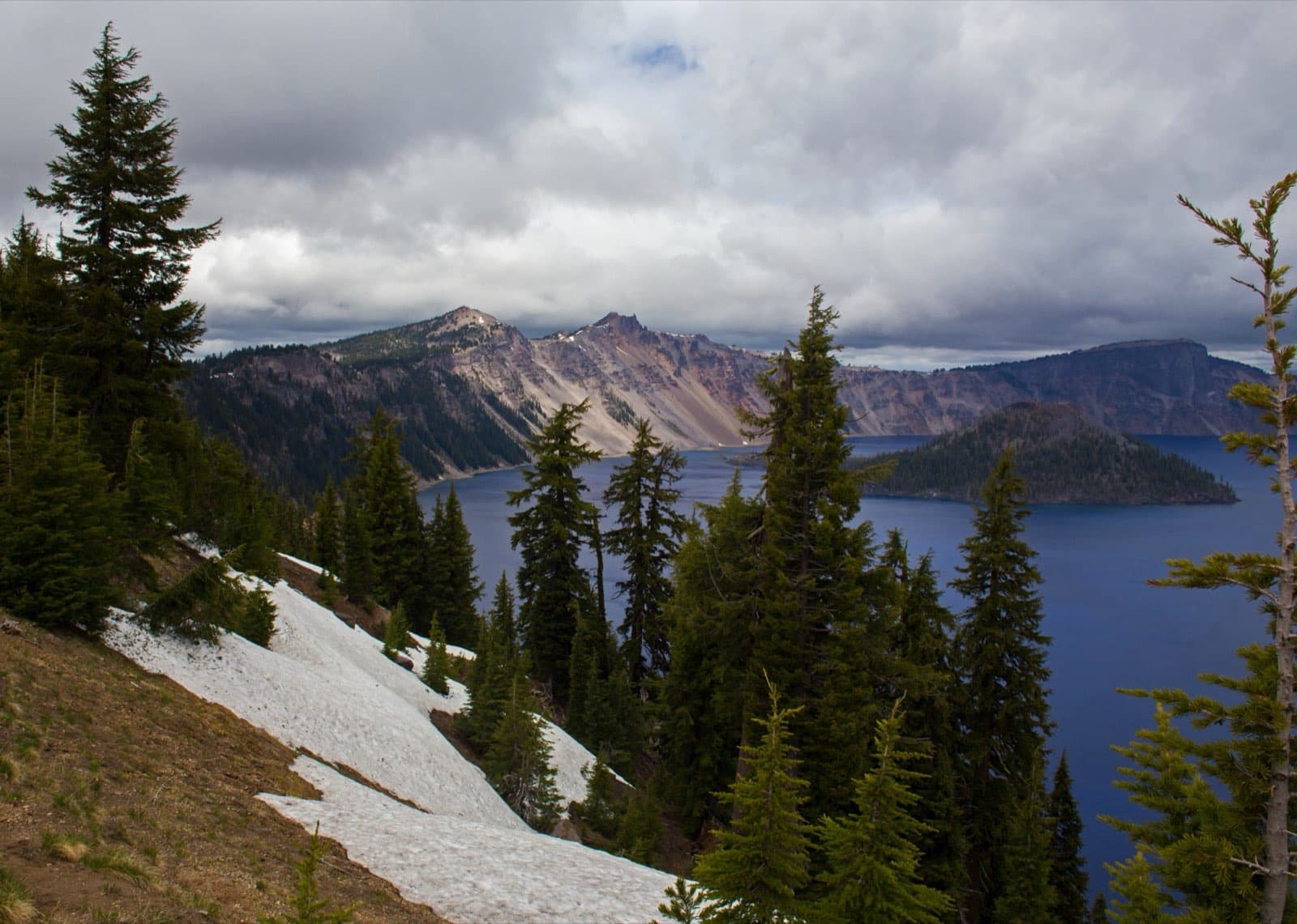 The deep blue water of Crater Lake under a clear sky, with the forested cinder cone of Wizard Island rising from the caldera and steep rim cliffs in the background, Crater Lake National Park, Oregon