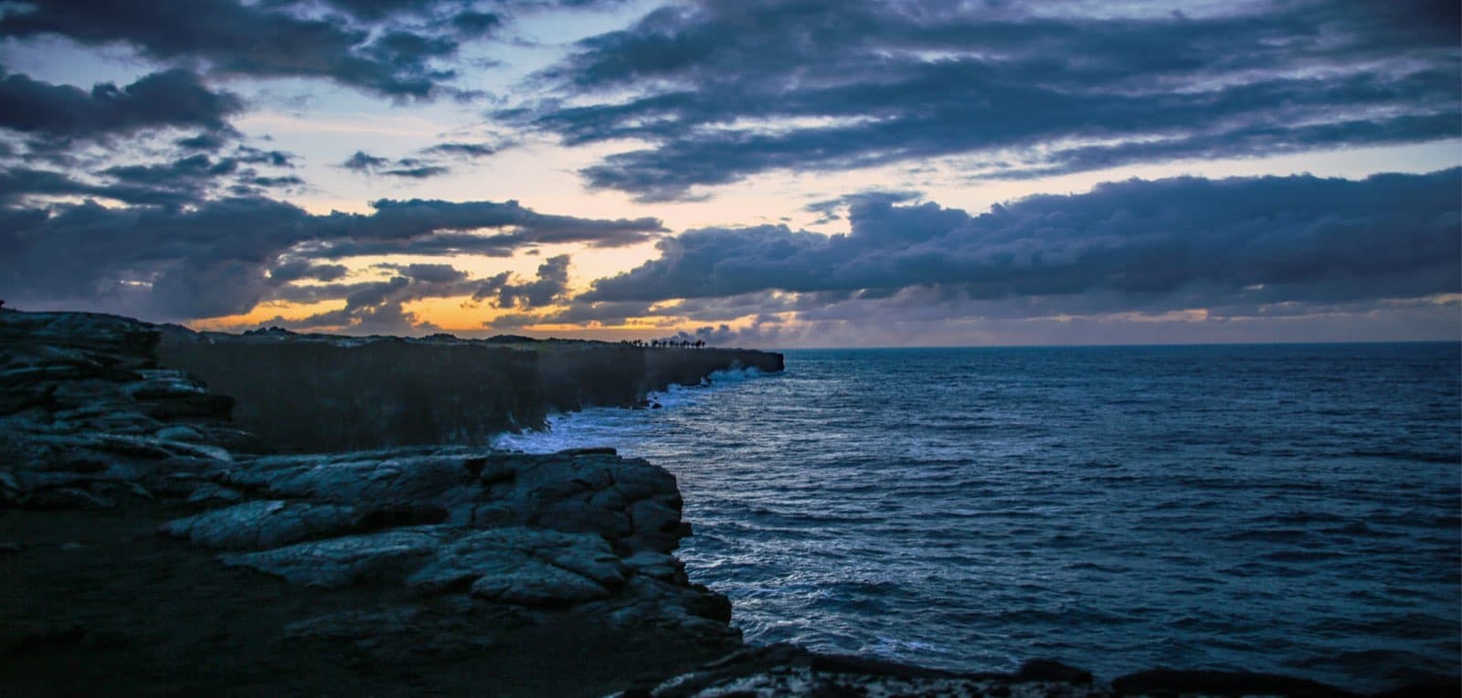 A dramatic wide dawn landscape at Hawaii Volcanoes National Park, with dark volcanic coastal terrain stretching beneath a deep early-morning sky.