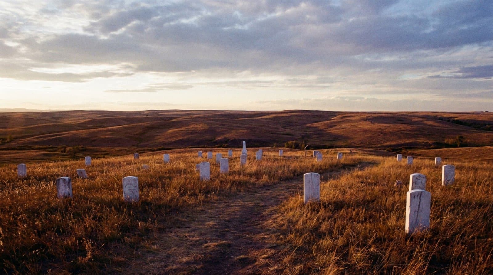 Last Stand Hill at Little Bighorn, scattered white marble markers across golden open prairie grass, big Montana sky with dramatic clouds, low warm evening light