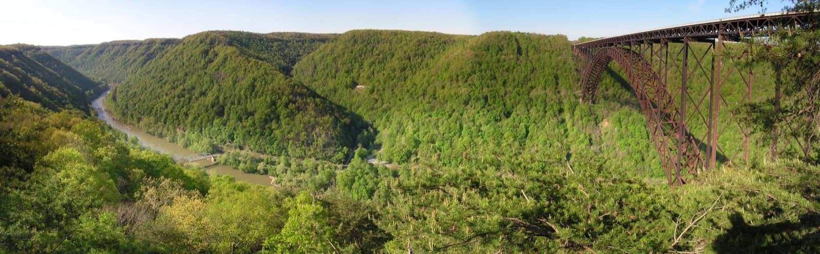 Wide panoramic view of New River Gorge National Park and Preserve, with the New River winding through the forested gorge and the steel-arch New River Gorge Bridge spanning the canyon, seen from the National Park Service overlook near Fayetteville, West Virginia.