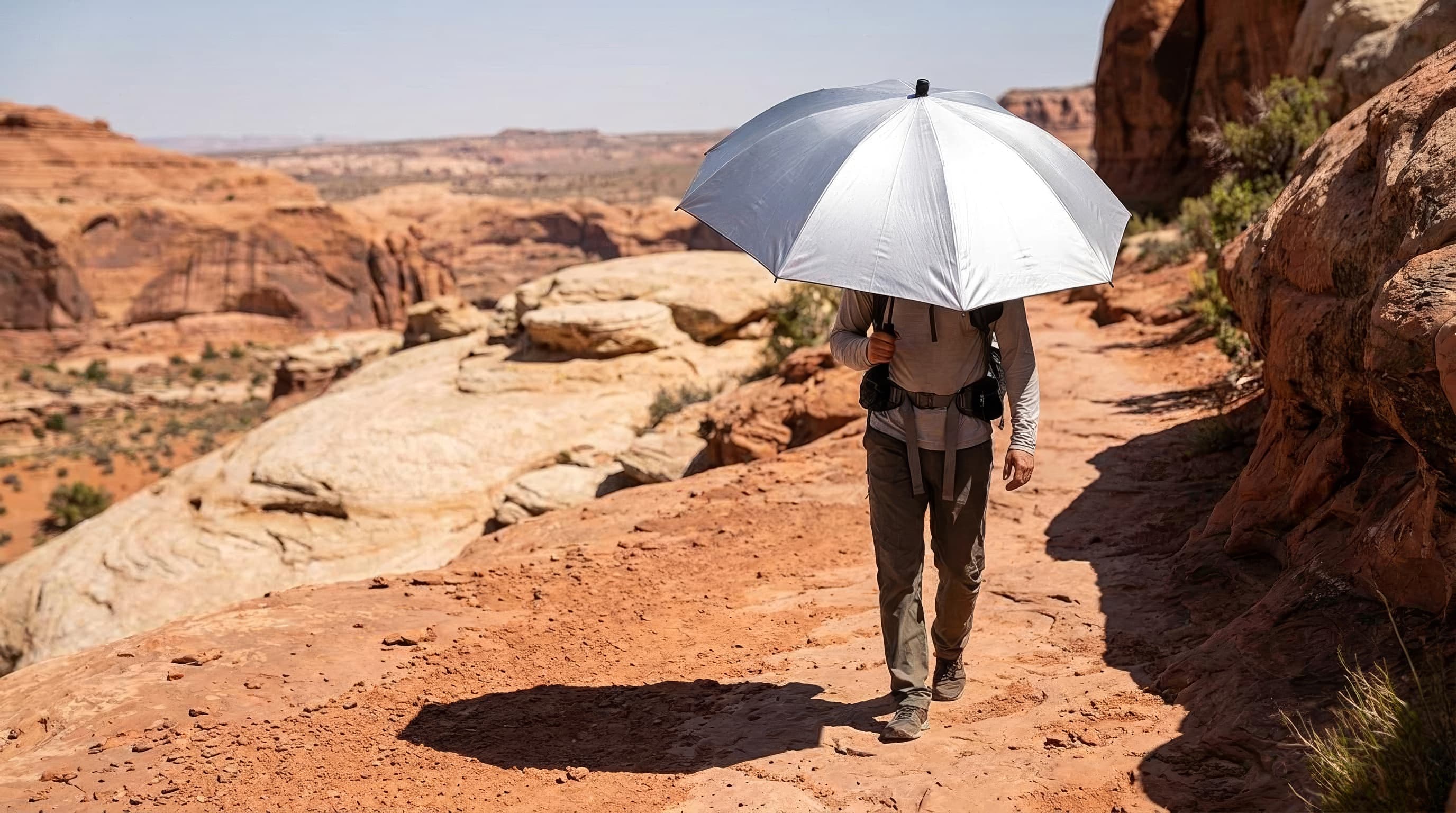 A hiker carrying a silver reflective trekking umbrella across an exposed desert trail