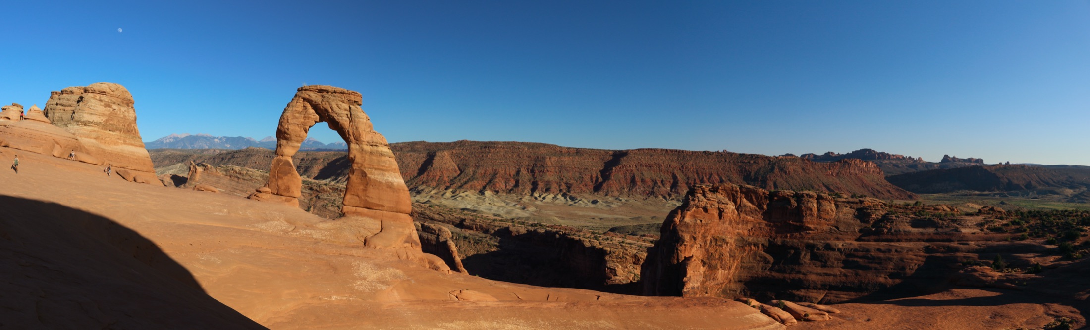 Panoramic view of Delicate Arch, the freestanding sandstone arch in Arches National Park, Utah, with the snow-capped La Sal Mountains in the distance under a clear sky