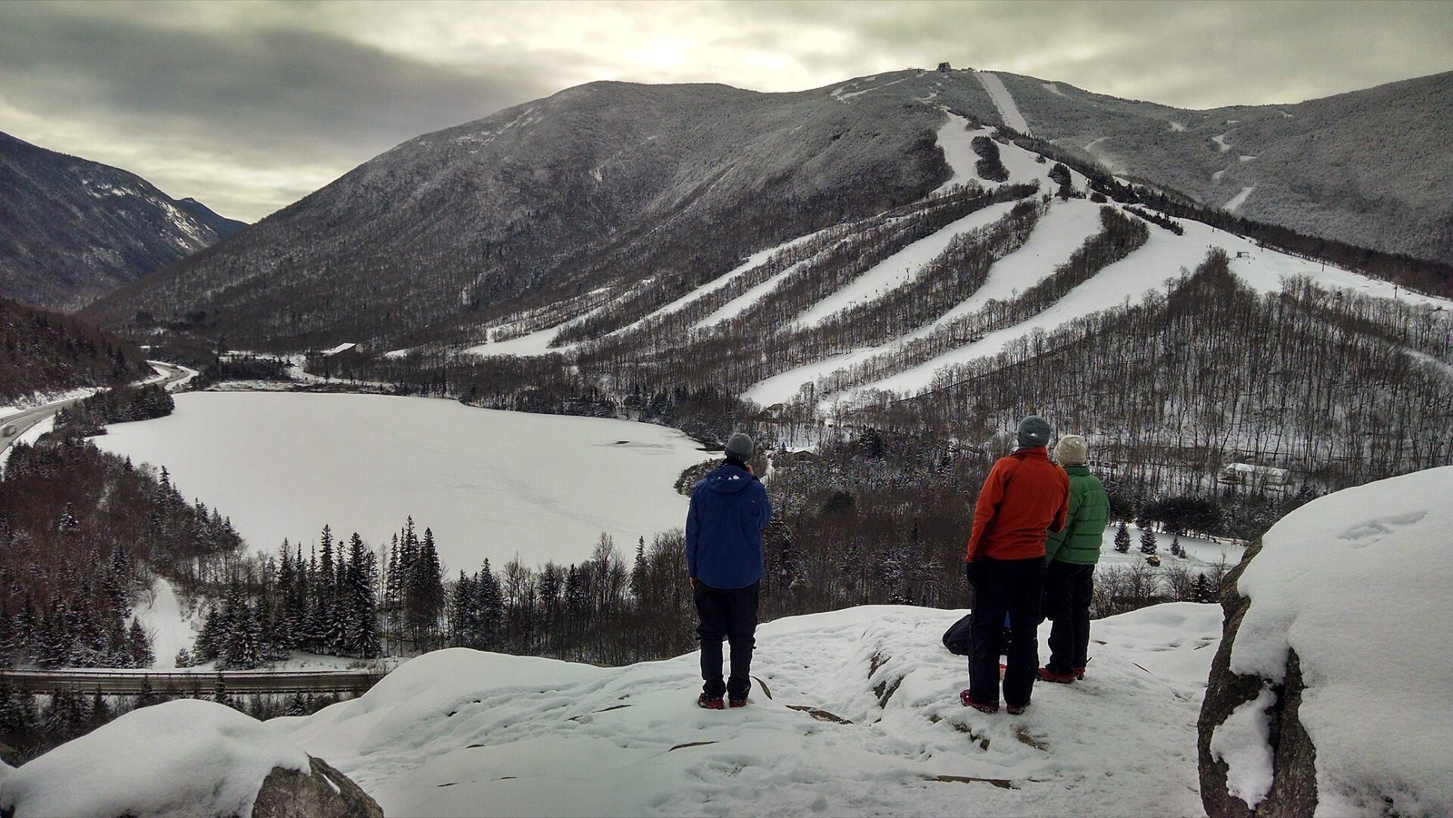 Three hikers on snowy Artists Bluff looking over frozen Echo Lake to the ski trails of Cannon Mountain