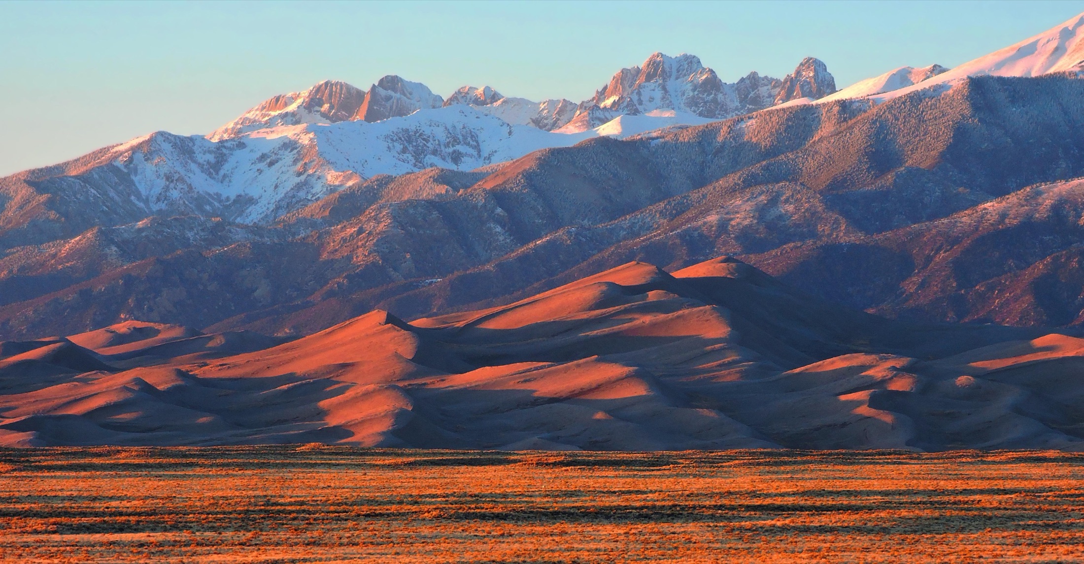 Warm alpenglow light on the rippled sand dunes of Great Sand Dunes National Park and Preserve, with the snow-dusted Sangre de Cristo Mountains rising behind them at sunset.