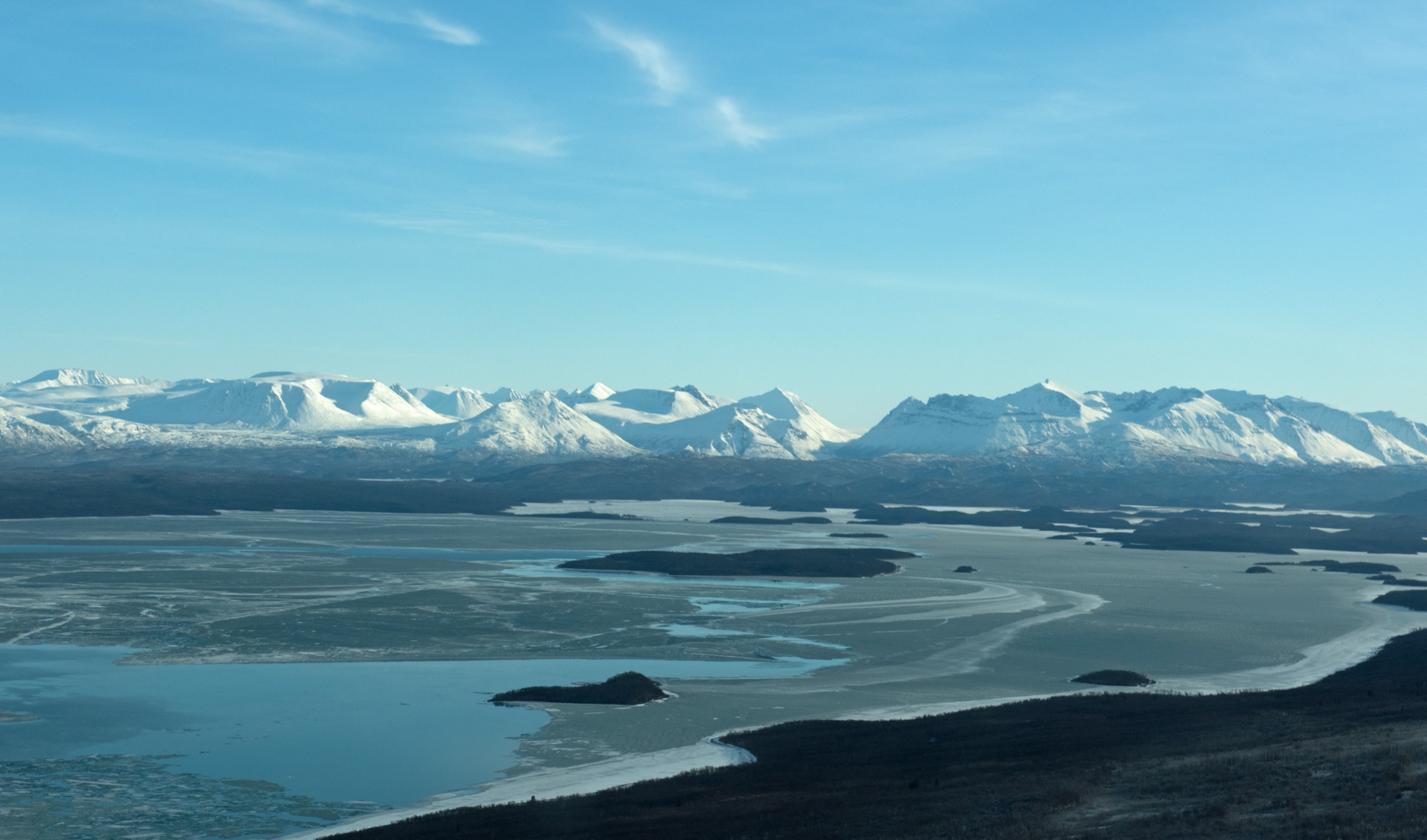 Snow-dusted shoreline and forested islands rising from the calm waters of the Bay of Islands on Naknek Lake in Katmai National Park and Preserve, Alaska, with low mountains in the distance under a soft winter sky.