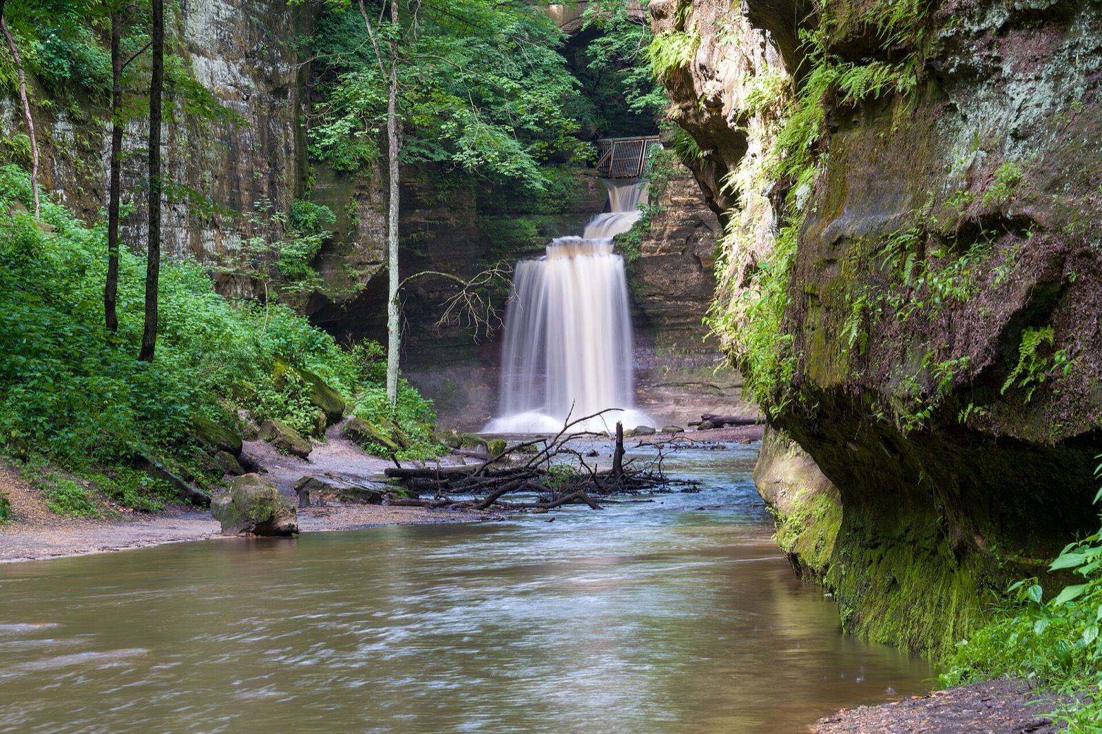 The Lower Dells waterfall at Matthiessen, a broad curtain of water below the footbridge, framed by moss-covered sandstone walls