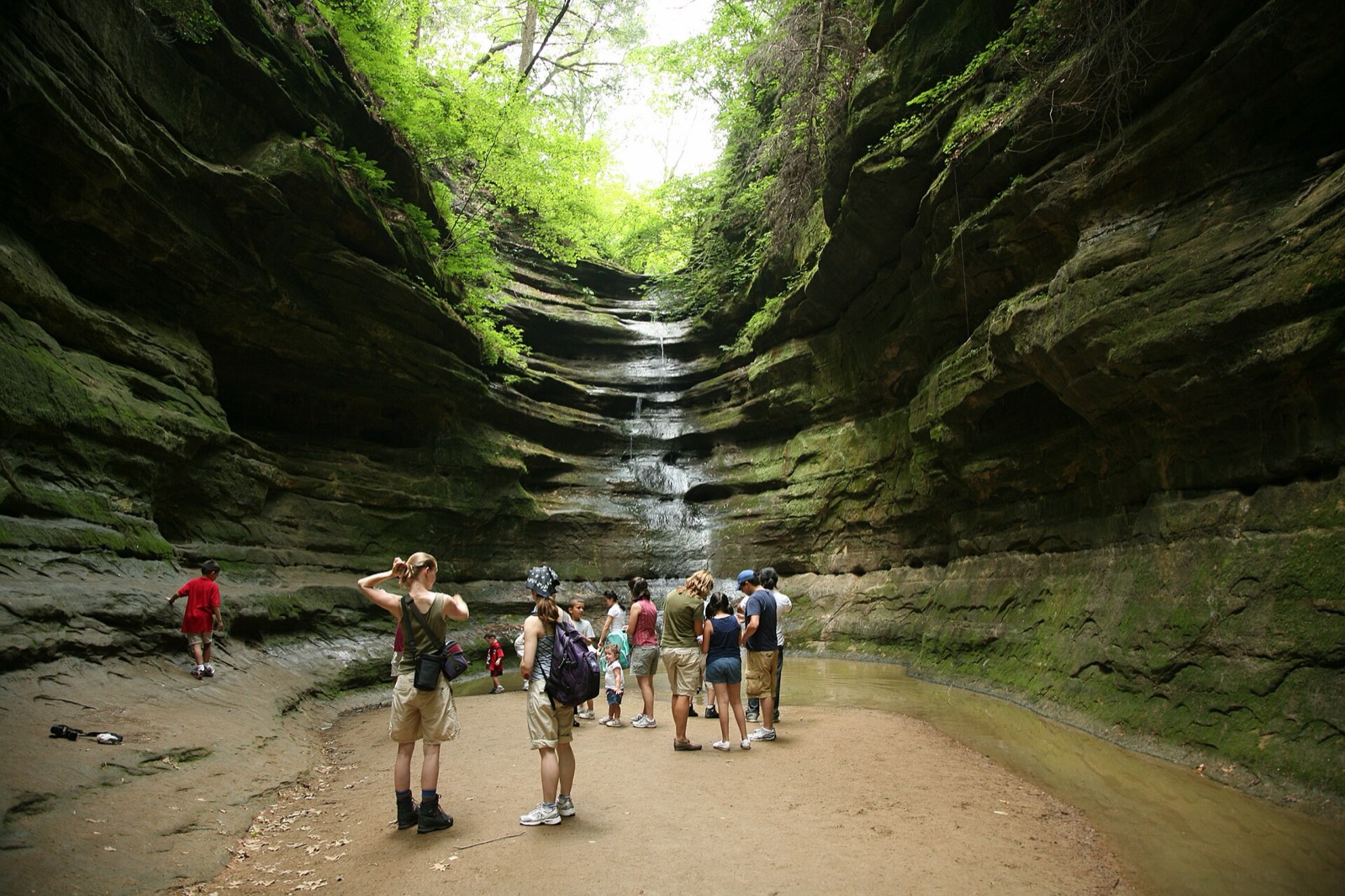 Hikers at the base of a sandstone box canyon at Starved Rock, its seasonal waterfall threading down the moss-lined wall